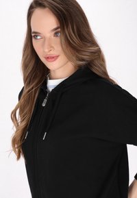 Schwarze Zip-up-Hoodie mit Kordelzügen, aus weichem Material. Über einem weiß-blauen gestreiften Shirt getragen. Nahaufnahme des Halsausschnitts und der Kapuze.