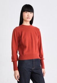 Roter Strickpullover mit gerippten Akzenten, Rundhalsausschnitt und langen Ärmeln, kombiniert mit dunklen Denim-Hosen. Glatte Textur, lässige Passform.