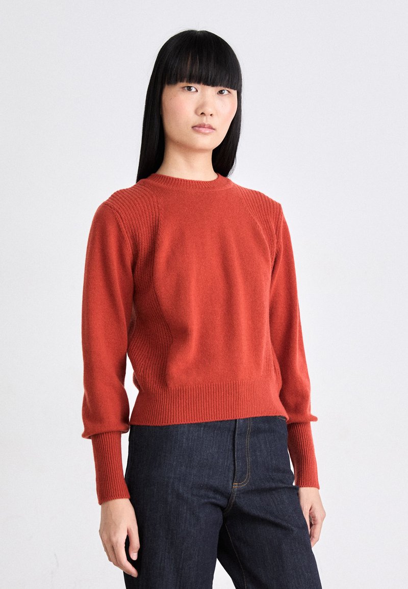 Roter Strickpullover mit gerippten Akzenten, Rundhalsausschnitt und langen Ärmeln, kombiniert mit dunklen Denim-Hosen. Glatte Textur, lässige Passform.