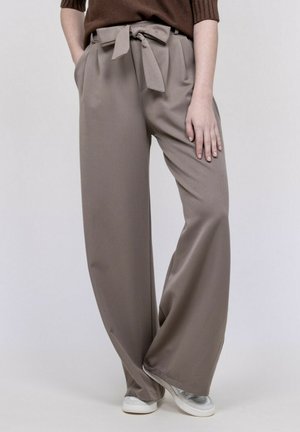 Femme portant un pantalon large taupe à ceinture nouée devant, des baskets blanches, et un pull côtelé marron, les mains dans les poches.