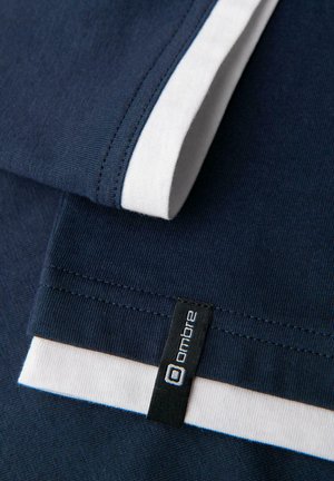 Meriininsininen kangas valkoisilla yksityiskohdilla hihoissa, mukana näkyvä musta etiketti, jossa on valkoista tekstiä. Siloinen pinta, rento muotoilu.