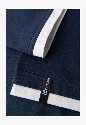 Granatowy materiał z białymi akcentami przy rękawach, z widoczną czarną metką z białym tekstem. Gładka tekstura, casualowy design.