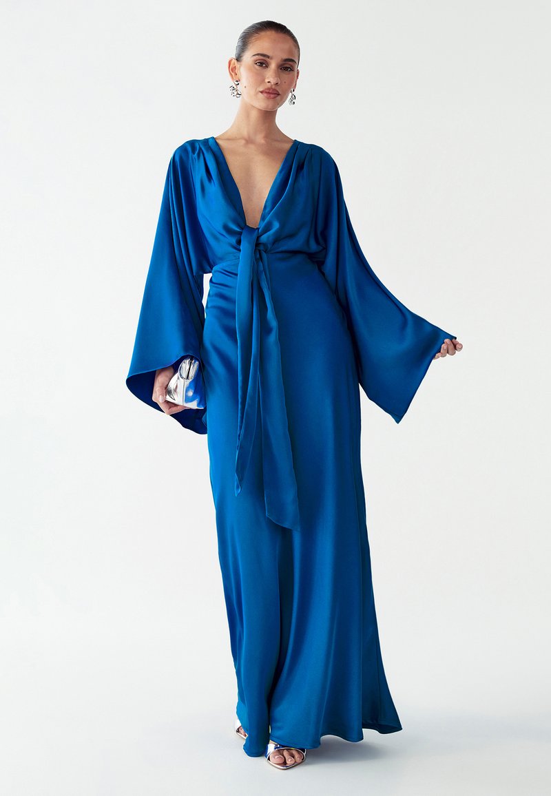 Lange, vloeiende blauwe satijnen jurk met wijde mouwen, diepe V-hals, tailleband en een silhouet tot op de vloer. Metalen clutch en open teenschoenen.
