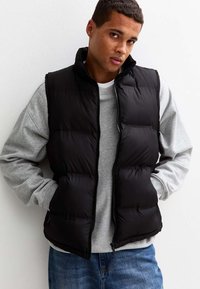 Czarna kamizelka puchowa z zapięciem na zamek, bez rękawów, o pikowanym wzorze, nałożona na szary sweter, w zestawie z niebieskimi dżinsami.