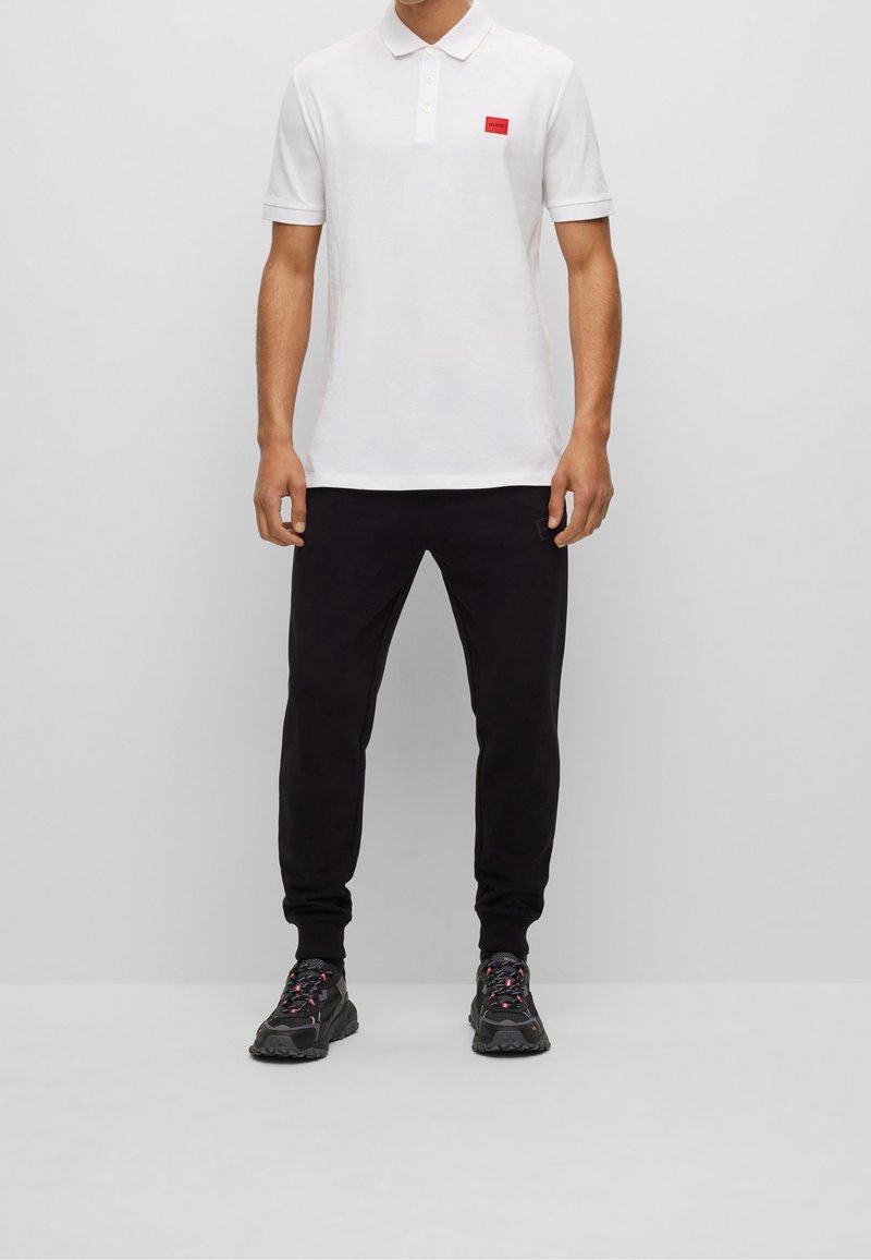 Homme portant un polo blanc avec un petit logo rouge sur la poitrine, un pantalon de jogging noir et des chaussures de sport noires avec des accents roses.
