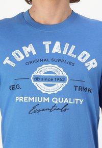 Zils kokvilnas T-krekls ar apaļu kaklu, īsām piedurknēm un baltiem drukātiem tekstiem un logotipu, tostarp "Tom Tailor" un "Since 1962."