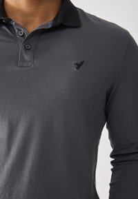 Long-sleeve polo shirt in dark gray cotton with a black collar. Features a subtle embroidered logo on the chest. Smooth texture.