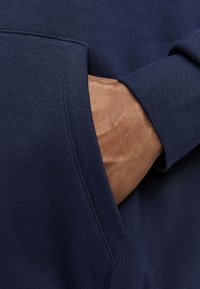 Felpa blu navy in tessuto testurizzato, con tasca laterale e polsini a costine. In primo piano si vede una mano appoggiata nella tasca.