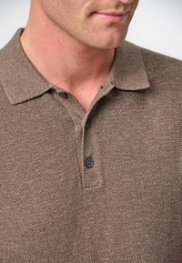 Brun texturerad pikétröja med krage och två svarta knappar vid knäppningen. Tyget har ett vävt utseende.