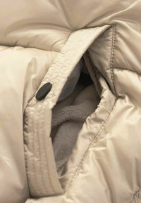Gros plan d'une poche de veste matelassée beige avec un bouton-pression noir et une doublure en polaire douce à l'intérieur de la poche.