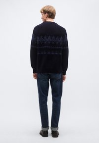 Sötétkék kötött pulóver geometrikus mintákkal kék árnyalatokban, sötét farmerrel és barna cipővel párosítva, hátulnézetben.