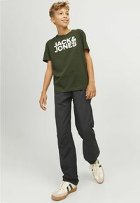 Zaļš kokvilnas T-krekls ar baltiem burtiem "JACK & JONES", apvienots ar melnām biksēm un beežiem sporta apaviem ar zaļām svītrām.