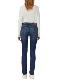 Jeans en denim bleu foncé avec une coupe slim, cinq poches et une patch en cuir marron sur la taille, associés à un haut à manches longues léger.