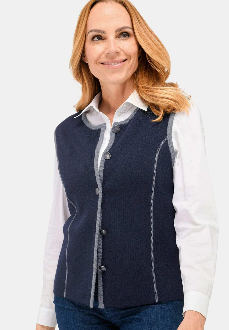 Dunkelblau gestrickte Weste mit rundem Ausschnitt, sechs dunklen Knöpfen, grauen Nähten entlang der Kanten und taillierter Passform über einem weißen Hemd.