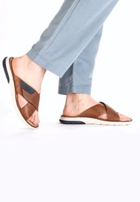 Braune Ledersandalen mit einem strukturierten Kreuzriemen-Design. Flexible weiße Sohle mit einem schwarzen Akzent. Getragen zu hellblauen Hosen.