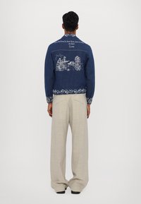 Denim jakna z modrim materialom, zapleteno belo vezenje na hrbtni strani, ki prikazuje prizore, ter okrašeni manšeti; v kombinaciji z svetlo bež hlačami.