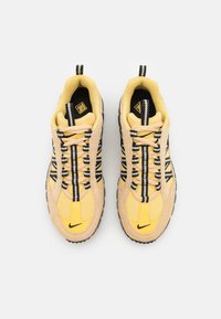Baskets jaune et beige avec une tige en tissu texturé, des lacets rayés, des accents noirs et une semelle extérieure en caoutchouc. Logo Nike sur le côté.