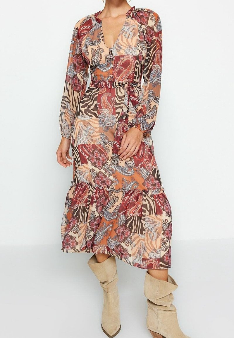 Femme portant une robe midi patchwork à manches longues avec imprimé floral et animal, ceinture nouée à la taille et bottes beige décontractées.