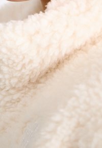 Sweter w kolorze kremowym przypominający wełnę, o miękkiej, teksturowanej, puszystej powierzchni; ukazuje zbliżenie na przytulny, pętelkowy materiał.