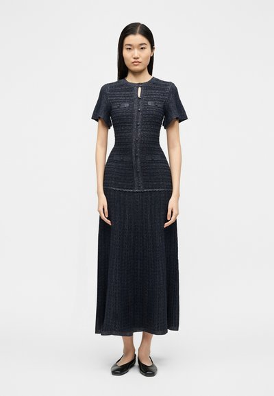 Γυναίκα στέκεται φορώντας ένα πουλόβερ φόρεμα σκούρου μπλε χρώματος με υφή, κοντά μανίκια, κουμπιά μπροστά, λαιμόκοψη με άνοιγμα και μαύρα παπούτσια slip-on.