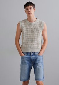 Top bej, tricotat, fără mâneci, cu un model texturat, combinat cu șorturi din denim ușor albastre. Modelul stă cu mâinile în buzunare.