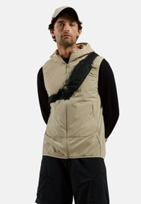 Beige padded vest with hood, zip closure, and two side pockets. Black crossbody bag worn over the shoulder; model also wears black shorts.