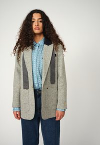 Light gray wool blazer with a textured finish, contrasting dark gray accents on the lapels, paired with a blue patterned shirt and dark denim.