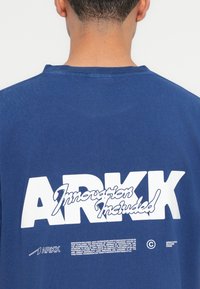 Plava pamučna majica s velikim bijelim logom "ARKK" i natpisom "Inovacija uključena" na stražnjoj strani. Dizajn s okruglim izrezom i opuštenim krojem.