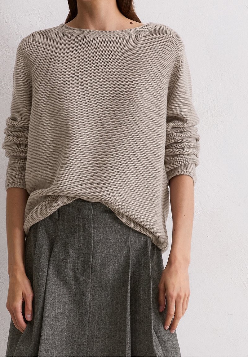 Femme portant un pull en tricot beige aux manches retroussées, rentré dans une jupe plissée grise, debout contre un mur blanc.