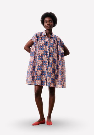 Femme noire debout, les mains derrière le dos, portant une robe ample au genou avec des motifs géométriques bleus, rouges et beiges, et des chaussures plates rouges.