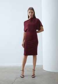 Burgundijas krāsas ceļgalu garuma kleita ar vienu plecu, izgatavota no gludas auduma, cieši pieguļ ķermenim; kombinēta ar melnām sandālēm ar siksniņām.