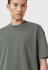 Olivengrøn bomulds t-shirt med rund halsudskæring, korte ærmer og lille broderet branding på venstre bryst, som har en glat tekstur.