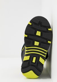 Schwarze Gummisohle mit ausgeprägtem Profil, versehen mit gelben Akzenten und strukturierten Mustern. Sichtbare Größenmarkierung und strukturelle Details.