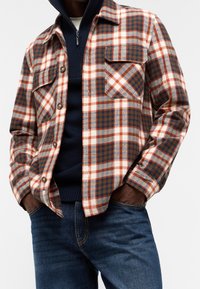 Kostkovaná flanelová košile v oranžové, modré a bílé barvě, s knoflíkovou légu vpředu, dvěma náprsními kapsami a volnějším střihem, v kombinaci s tmavými džíny.
