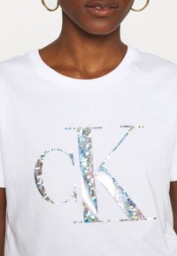 Camiseta de algodón blanca con un gran logotipo holográfico "CK" que brilla. Cuello redondo y mangas cortas.
