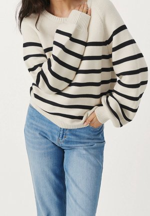 Donna che indossa un maglione crema con strisce orizzontali nere e jeans azzurri, una mano in tasca e l'altra che tocca il colletto.