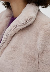 Faux fur jakke i lys beige med en blød tekstur. Har en opretstående krave og en lynlås med dekorative perler langs kanten.