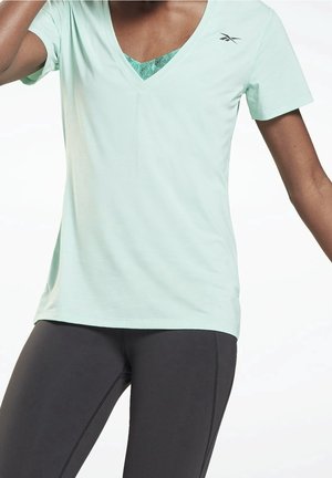 Vrouw draagt een lichtgroen Reebok V-hals T-shirt en zwarte leggings, poserend met één arm omhoog en één arm ontspannen.