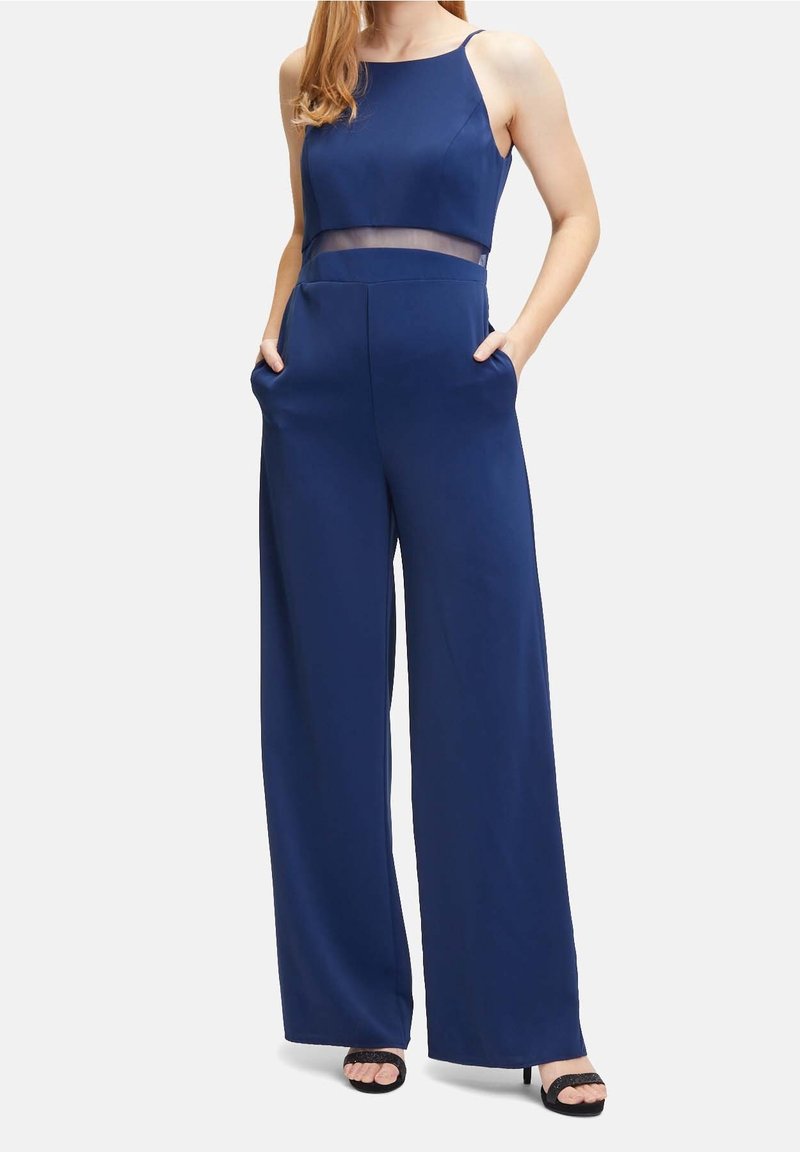 Marinblå jumpsuit med ärmlös design, genomskinlig midja, vida ben, sidfickor och slät textur. Bärs med svarta högklackade sandaler.