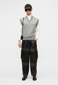 Γκρι γιλέκο με σχέδια πάνω από λευκό πουκάμισο με μακριά μανίκια, συνδυασμένο με μαύρο και καφέ παντελόνι cargo με τσέπες και φαρδιά εφαρμογή. Μαύρα γυαλιά ηλίου.