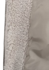 Lähikuva beige-värisestä fleecestä, jonka vieressä on sileä vaaleanbeige kangas ja hopeinen vetoketju kulkee reunan myötäisesti.