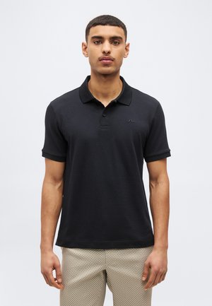 Mladý muž oblečený v čiernej polo košeli s krátkymi rukávmi a béžových nohaviciach so vzorom, stojaci pred jednofarebným svetlosivým pozadím.