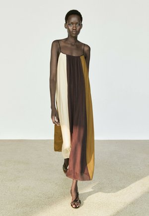 Frau geht, trägt ein ärmelloses Kleid mit Farbblock-Design mit vertikalen Paneelen in Beige, Braun und Senf und schwarzen Riemchensandalen auf hellem Boden.