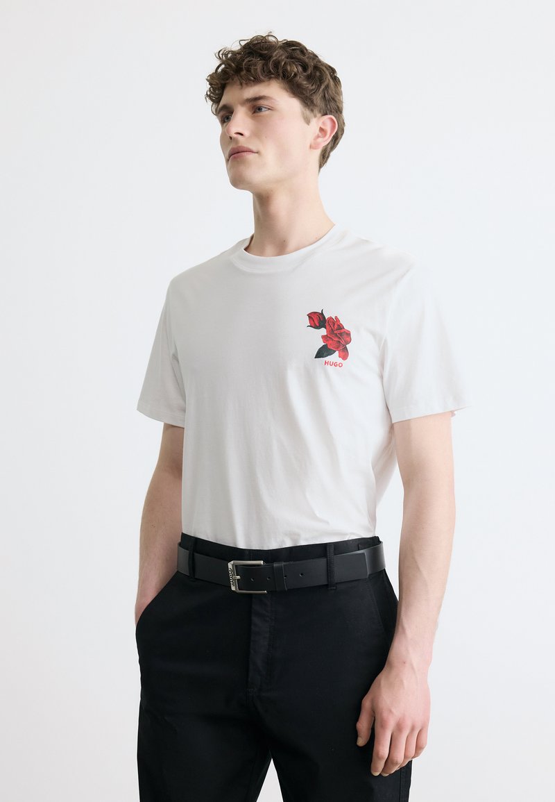 Νέος άνδρας που φοράει λευκό t-shirt με κόκκινο γραφικό τριαντάφυλλο και κείμενο «HUGO», μαύρο παντελόνι και μαύρη ζώνη, στέκεται μπροστά σε λευκό φόντο.