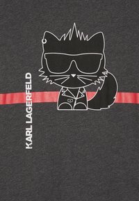 Kreskówkowy czarny kot w okularach przeciwsłonecznych i skórzanej kurtce, umieszczony nad czerwoną paskiem obok pionowego napisu "KARL LAGERFELD" na ciemnoszarej tkaninie.