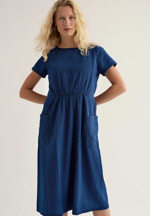 Szőke nő, kék farmer midi ruhában, rövid ujjakkal, elasztikus derékkal és elülső foltzsebekkel, kezeit a hátája mögött tartva áll.