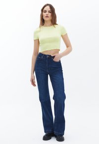 Gul cropped top med korte ærmer, parret med mørkeblå høj talje flared jeans. Model bærer sorte kraftige støvler. Hvid baggrund.