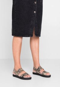 Tas sandalias rata-rata dengan desain cetakan ular, dilengkapi dengan dua tali lebar dan sol hitam. Tali tersebut terbuat dari material dengan tekstur.