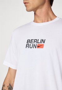 Man wearing white T-shirt with "BERLIN RUN" text and red Nike logo on chest, showing tattooed forearm and side profile of face.