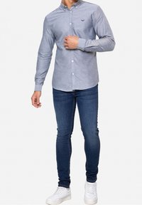 Chemise grise boutonnée avec un motif texturé, associée à un jean slim bleu foncé et des baskets blanches. Comprend un petit logo brodé.